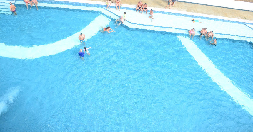 Piscina de água azul vista do alto, com hóspedes sentados na borda e brincando na água